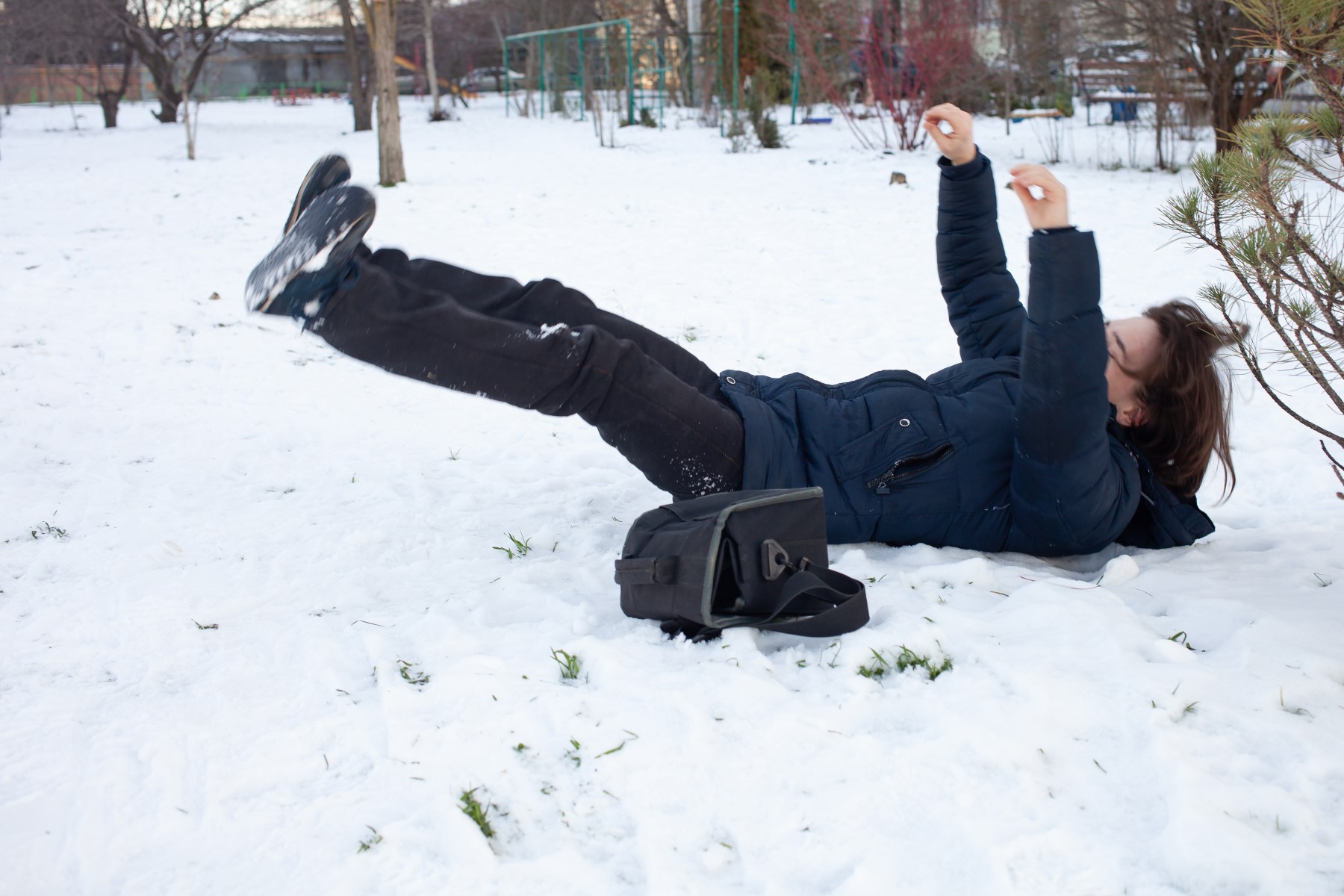 A person slipping and falling on the snow in a park during winter, with a black bag next to them, emphasizing the hazards of icy conditions