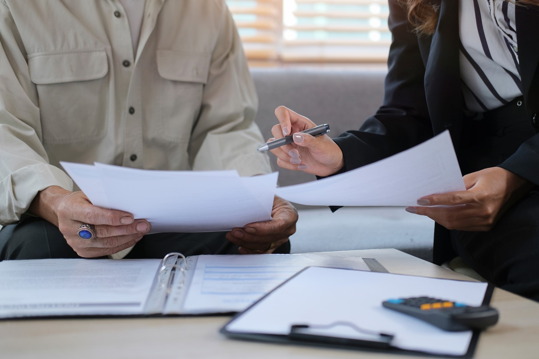 Close-up of two people reviewing financial documents, one person holding a pen and the other holding papers, during a discussion, highlighting a professional setting.