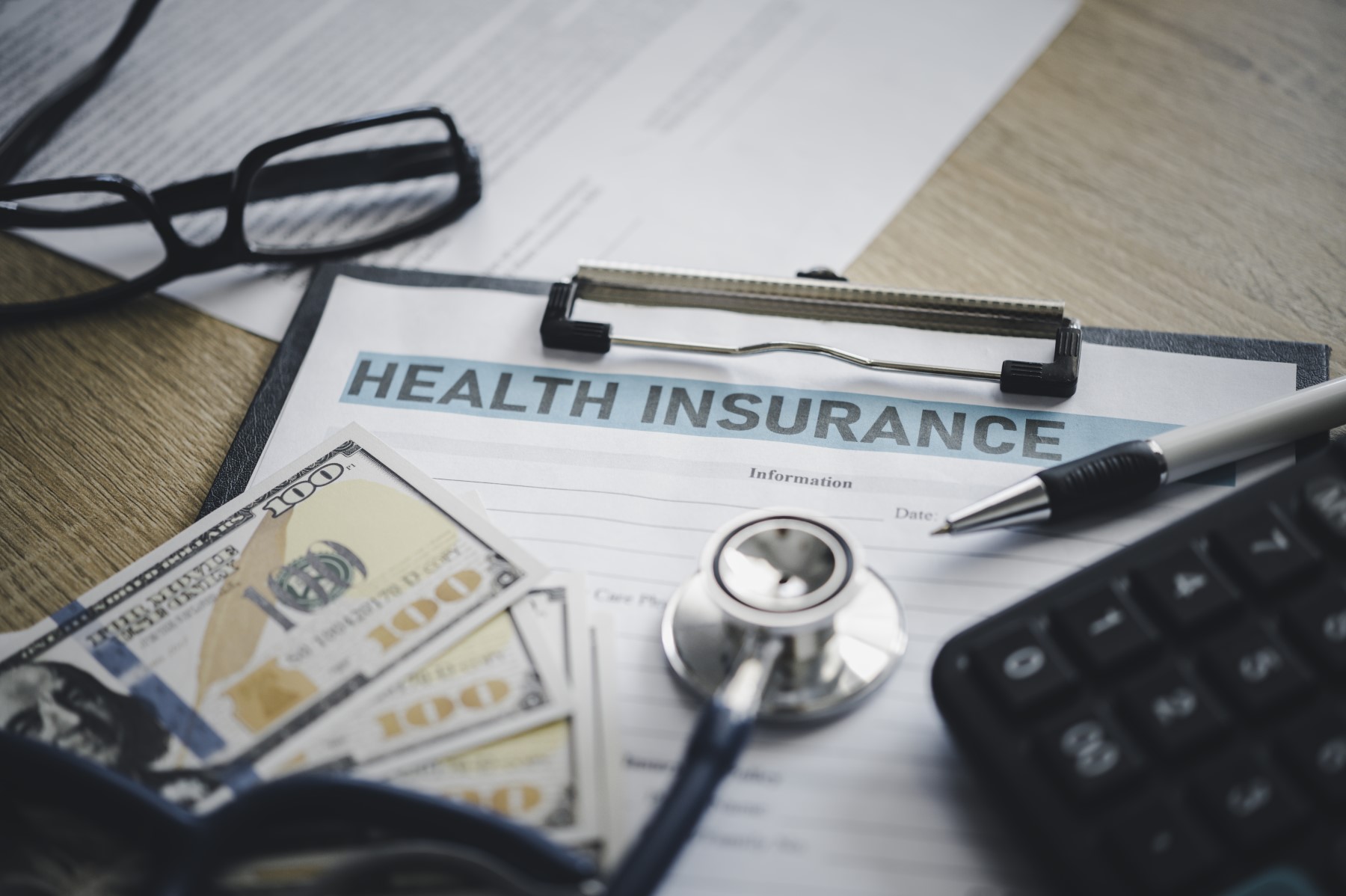 Close-up of a health insurance form on a clipboard surrounded by a stethoscope, cash, a pen, calculator, and eyeglasses, symbolizing the financial aspects of healthcare and insurance.