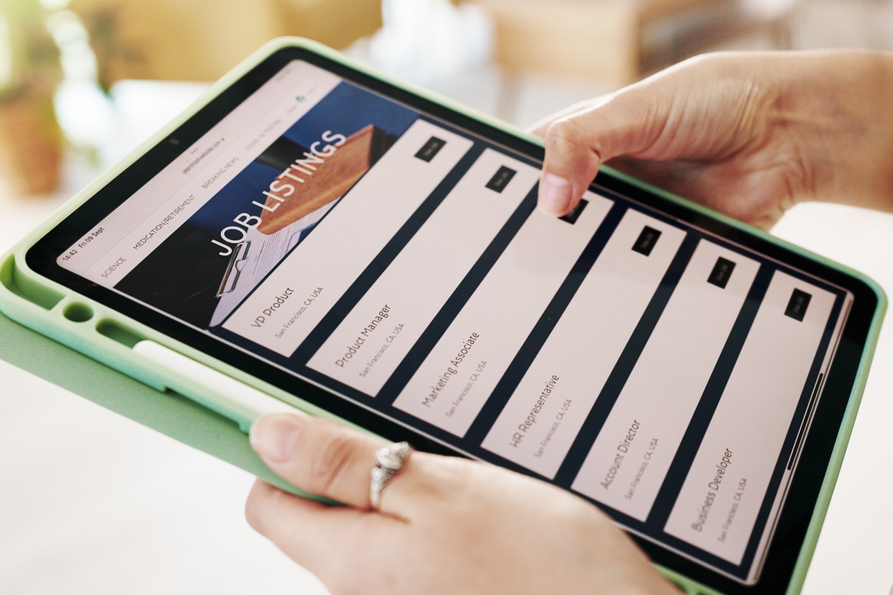 A close-up image of a person holding a green-cased tablet displaying a "Job Listings" webpage, with various job roles like "VP Product" and "Marketing Associate" visible on the screen. The person is using their right hand to scroll through the listings.