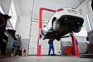 Car on a hydraulic lift in an auto repair shop while a mechanic in blue overalls works underneath the vehicle during maintenance.