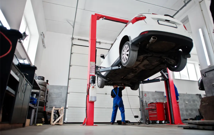 Car on a hydraulic lift in an auto repair shop while a mechanic in blue overalls works underneath the vehicle during maintenance.