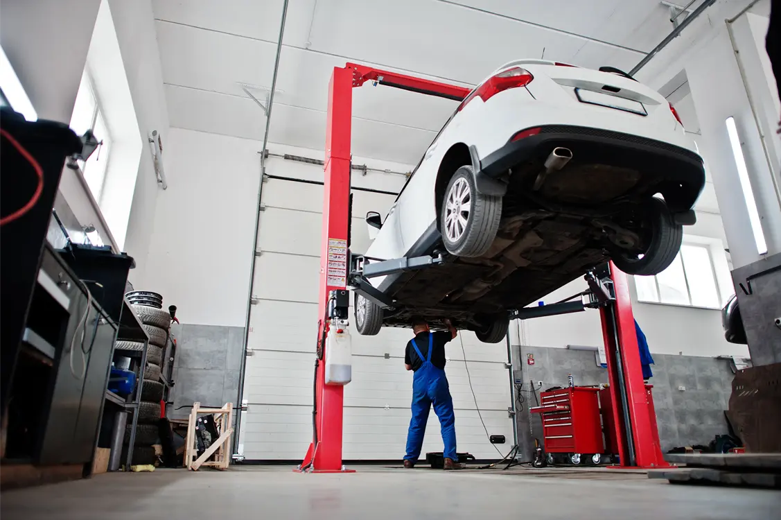 Car on a hydraulic lift in an auto repair shop while a mechanic in blue overalls works underneath the vehicle during maintenance.